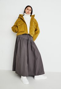 Żółta krótka kurtka na biały golf, w połączeniu z długą szaro-skalą spódnicą i białymi trampkami. Otoczenie jest minimalistyczne i jasne.