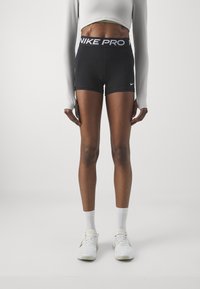Svarta Nike Pro-short med en bred vit midja. Modellen har på sig en åtsittande vit långärmad tröja och vita träningsskor med gula detaljer.
