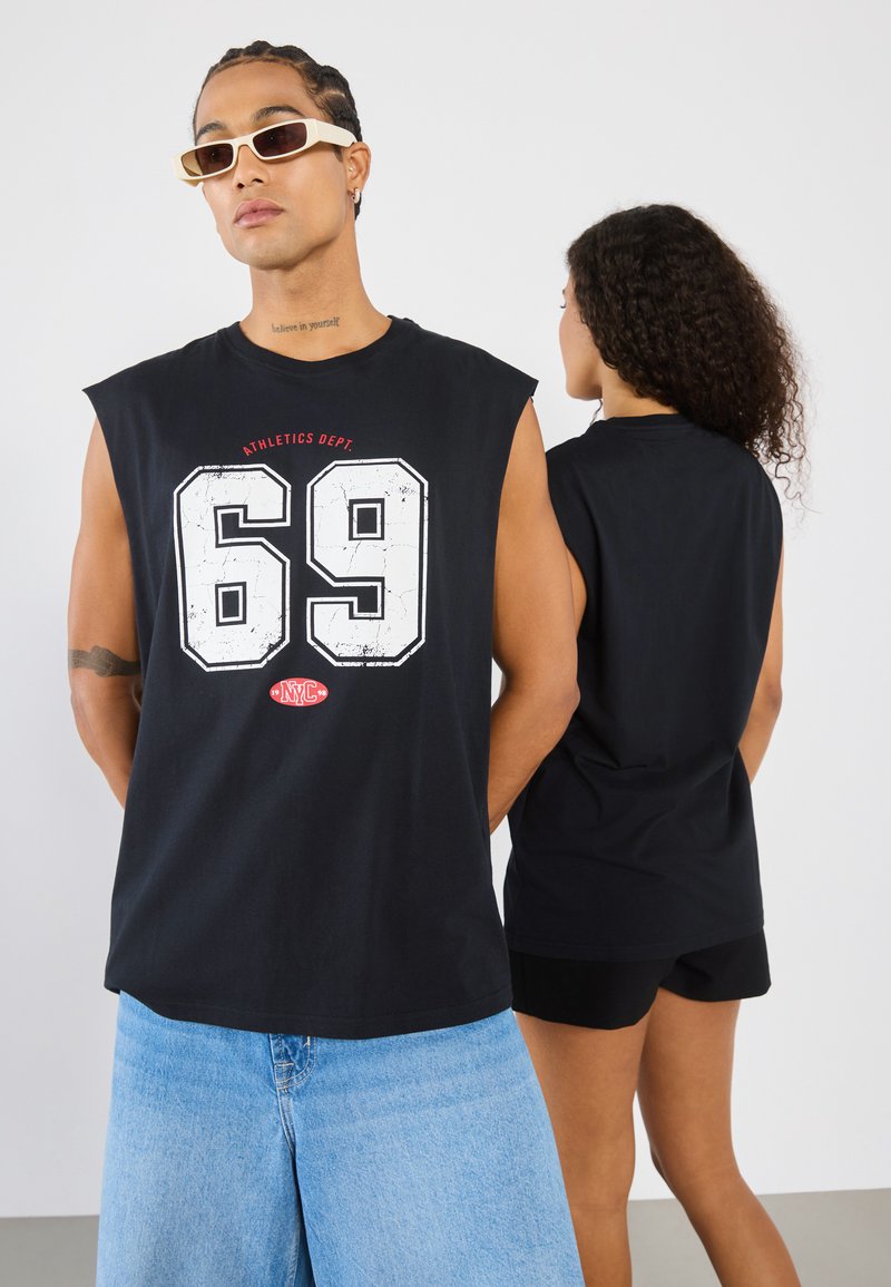 Svart ermeløs t-skjorte med en stor hvit grafikk "69" og teksten "ATHLETICS DEPT.", kombinert med lyseblå denimshorts og solbriller.