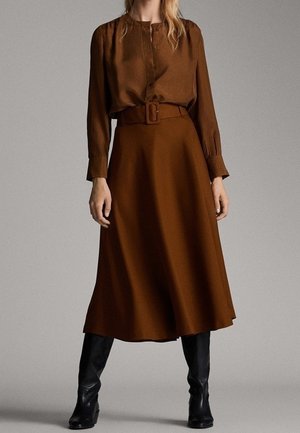 Vrouw draagt een bruine blouse met lange mouwen, ingestopt in een bruine midirok met een bijpassende riem en zwarte kniehoge laarzen.
