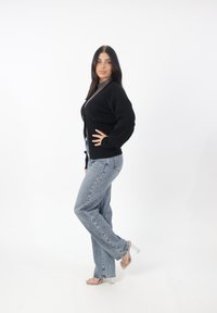 Schwarze Strickjacke, graues Oberteil, hellblaue Jeans mit ausgestelltem Design und transparente High-Heelsandalen, auf einem weißen Hintergrund posiert.