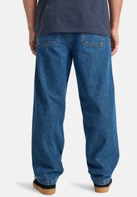 Pantalones de mezclilla azul con un corte holgado, cuentan con bolsillos traseros, costuras visibles y una textura ligeramente desgastada. Combinados con zapatos marrones.