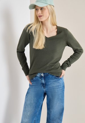 Femme blonde portant une casquette vert menthe, une chemise à manches longues vert foncé et un jean bleu, debout les mains dans les poches, regardant vers la droite.