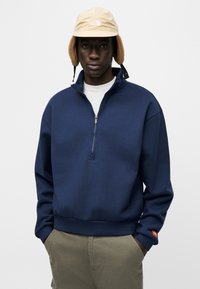 Πολύ απαλή μπλούζα μπλε ναυτικού με ψηλό γιακά, ημικαμπαρντίνα και ριμπ τελείωμα. Χακί παντελόνι με πλαϊνές τσέπες. Μπεζ καπέλο με καλύμματα αυτιών και λογότυπο.