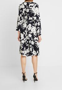 Floral dress with a navy background, featuring white flowers. Long sleeves, cinched waist, and calf-length with a slit at the back. Black heels.