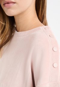 Sudadera rosa claro acanalada con cuello redondo y botones decorativos a lo largo de la costura del hombro, con una textura suave.