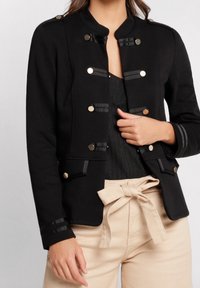 Zwarte jack met staande kraag, gouden knopen en gestreepte accenten, gecombineerd met lichtgekleurde broek met riem.