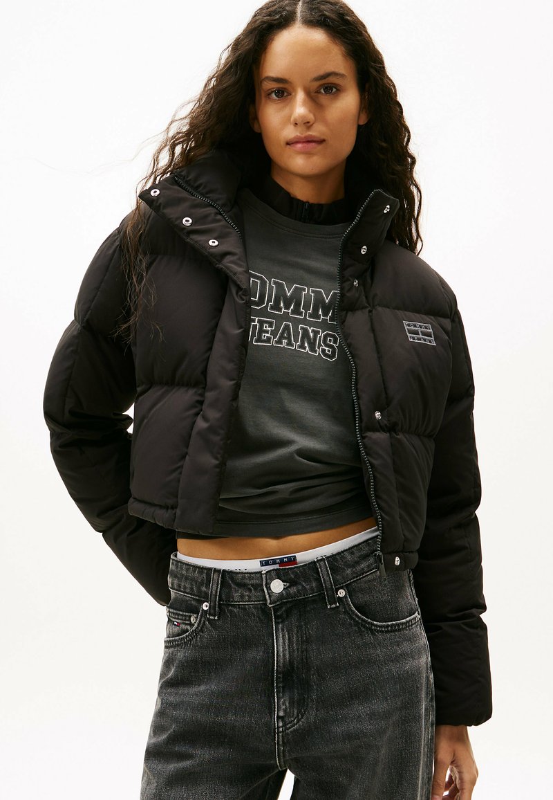 Svart kort pufferjacka med stor krage, frontknäppning och logotypmärke. Bärs över en mörkgrå skjorta och urtvättade svarta jeans.