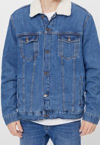 Muž v modré džínové bundě s bílým fleecovým límcem a modrých džínách, stojící před jednobarevným pozadím.