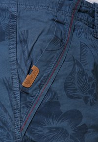 Blue shorts featuring a floral pattern, zipper closure with red stitching, and a tan leather tag with embossed text.