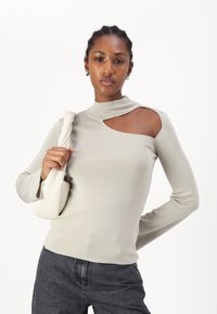 Haut à manches longues gris clair avec un col montant et un design découpé sur l'épaule, associé à un jean foncé et à un sac beige.
