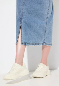 Valkoinen sneaker, jossa on teksturoitu pinta, pyöreä kärki ja paksu pohja. Yhdistetty vaaleansiniseen farkkuhameeseen, jossa on etureikä.