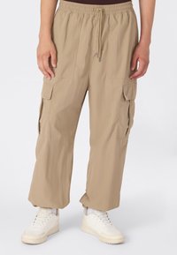 Luźne beżowe spodnie cargo z elastycznym paskiem i sznurkiem, w połączeniu z białymi sneakersami i białymi skarpetkami, noszone przez stojącą osobę.