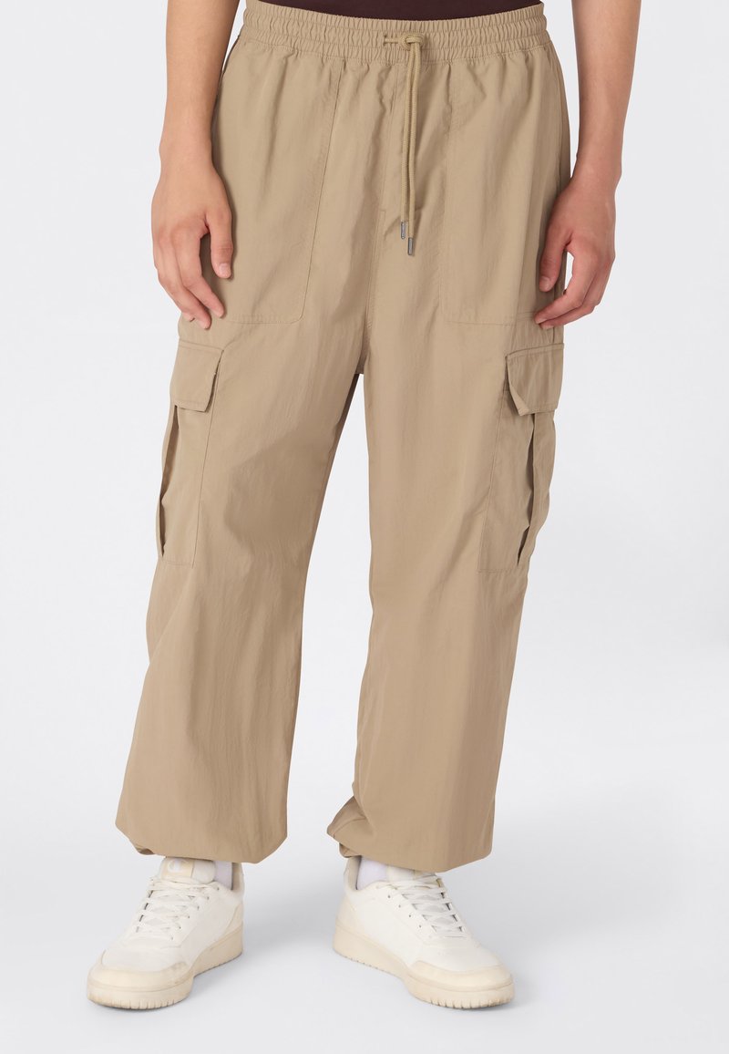 Luźne beżowe spodnie cargo z elastycznym paskiem i sznurkiem, w połączeniu z białymi sneakersami i białymi skarpetkami, noszone przez stojącą osobę.