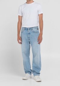 Man i en enkel vit t-shirt med korta ärmar, ljusblå löst sittande jeans och vita sneakers som står mot en enfärgad bakgrund.