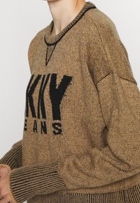 Pull marron en maille avec un logo noir. Il présente un col rond, des poignets côtelés et une texture épaisse. Mis en valeur par des surpiqûres contrastantes.