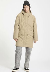 Cappotto lungo beige con cappuccio, tasche laterali e chiusura con zip. Indossato sopra pantaloni grigi con motivo e sneakers nere. Cuffia nera lavorata a maglia in testa.