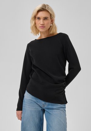 Žena s blond vlasmi má na sebe čierny top s dlhým rukávom a svetlomodré džínsy, stojac pred jednofarebným svetlosivým pozadím.