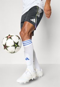 Shorts de football noirs avec des accents violets et verts, chaussettes blanches avec des rayures bleues, et chaussures blanches. Un ballon de football à motifs est à proximité.