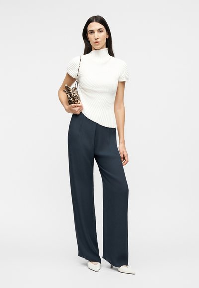 Γυναίκα που φοράει λευκό πλεκτό τοπ με κοντά μανίκια, μαύρο παντελόνι με φαρδιά μπατζάκια, λευκές γόβες, κρατώντας μικρή τσάντα ώμου με λεοπάρ σχέδιο.