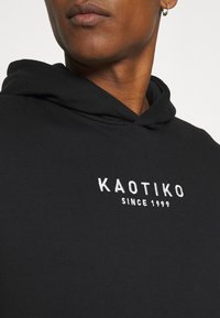 Ingrandimento di una persona che indossa un hoodie nero con la scritta bianca "KAOTIKO SINCE 1999" sul petto e un piccolo orecchino a cerchio nell'orecchio sinistro.