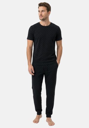 Mężczyzna stoi boso, ubrany w czarną koszulkę z krótkim rękawem i dopasowane czarne spodnie dresowe z delikatnym wzorem.