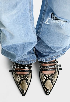 Buty z wzorem skóry węża o spiczastym czubku, z czarnymi paskami z podwójną klamrą i ćwiekami, noszone z jasnoniebieskimi, podartymi dżinsami.