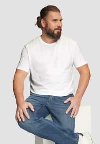 Weißes Baumwoll-T-Shirt mit kurzen Ärmeln und Rundhalsausschnitt, kombiniert mit hellblauen Jeans. Modell sitzt auf einem weißen Sockel.