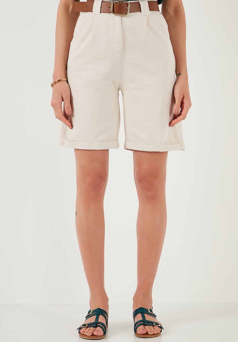 Pantaloni corti in cotone beige con orli arrotolati, caratterizzati da pieghe frontali e una cintura marrone con fibbia rettangolare. Indossati con sandali a strapiombo verdi scuri.