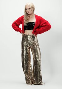 Czerwona dziana kardiganka, błyszczący czarny top typu crop i złote, cekinowe spodnie dzwony. Modelka ma na sobie metaliczne buty, a włosy ułożone w luźne fale.