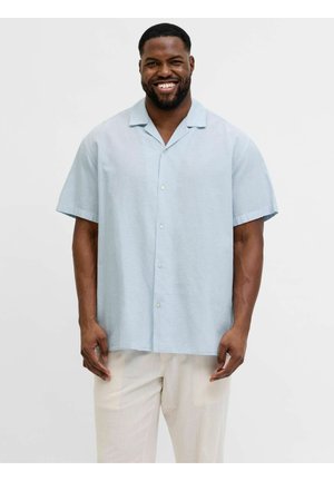 Mosolygó férfi világoskék rövid ujjú gombos ingben és világos színű nadrágban, egy egyszínű háttér előtt állva.