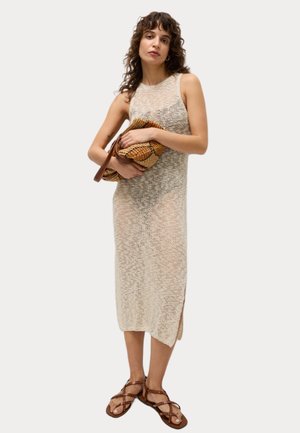 Vestido de punto ligero en crema con aberturas laterales, combinado con sandalias marrones con tiras y un clutch marrón texturizado. Cabello rizado y accesorios mínimos.