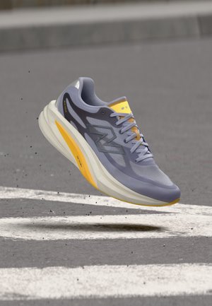 Chaussure de course gris clair et jaune avec une semelle épaisse flottant au-dessus d'une route aux marquages blancs.
