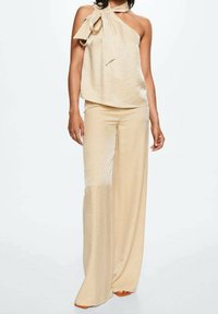 Débardeur beige sans manches avec un nœud sur une épaule, associé à un pantalon large assorti. Tissu lisse et brillant avec un léger éclat.