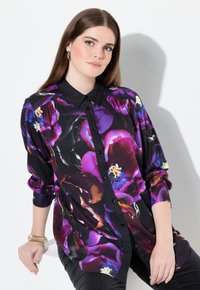 Ung kvinde med glat brunt hår iført en sort blomstret bluse med lilla, pink og orange blomster, siddende mod en hvid baggrund.
