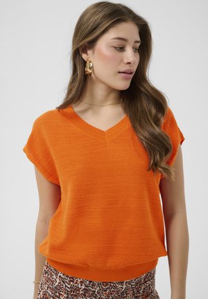 Femme aux longs cheveux bruns portant un pull à manches courtes col en V orange vif et une jupe à motifs, regardant sur le côté sur un fond uni.