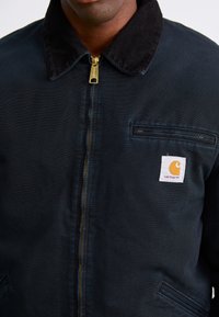 Nahaufnahme einer Person, die eine dunkelblaue Carhartt-Jacke mit einem schwarzen Cordkragen und einem goldenen Reißverschluss trägt. Das Markenlogo-Patch ist auf der Brust sichtbar.