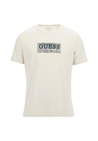 T-shirt color beige chiaro in cotone, con una grafica blu "GUESS EST. 1981" sulla parte anteriore e decorazioni a motivi geometrici. Design con scollo tondo.