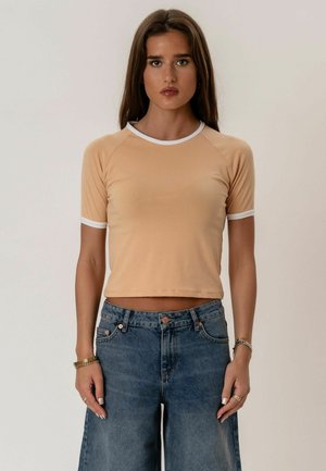 Młoda kobieta o długich brązowych włosach nosi beżowy krótki top z białym wykończeniem i wysokim stanem niebieskich dżinsów, stoi na jednolitym tle.