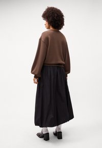 Barna, oversized pulóver, amelynek vállai ejtettek, fekete, rakott maxi szoknyával párosítva. A modellen szürke zoknik és fekete magassarkú cipők vannak.