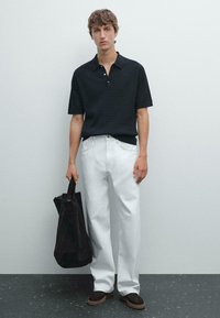 Polo shirt nero a maniche corte con un design testurizzato, abbinato a pantaloni bianchi a gamba larga, con una borsa a spalla in suede nero e scarpe marroni.