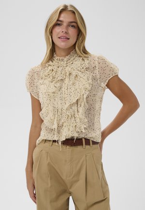 Beżowa bluzka z falbanianym kołnierzem i krótkimi rękawami. Posiada czarne groszki, lekką tkaninę i luźny krój. Połączona z beżowymi spodniami.