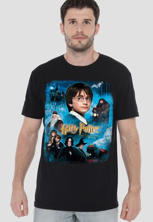 Junger Mann trägt ein schwarzes T-Shirt mit Harry-Potter-Charakteren und Schloss Hogwarts vor magischem blauem Hintergrund und goldenem Titeltext.