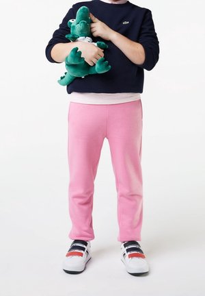 Lys Pink sweatpants med en glat tekstur; marineblå sweatshirt; hvide og sorte sko med røde accenter; grønt plysdinosaur legetøj i hånden.