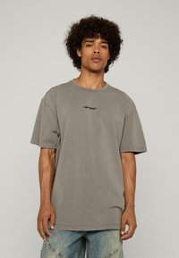 Jauns vīrietis ar cirtainiem matiem, tērpies bālganā pelēkā pārāk lielā T-kreklā ar uzrakstu "ZAUDĒTA JAUNĪBA" un gaiši zilās džinsa biksēs pret vienkāršu fona.