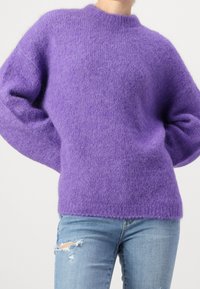 Person som bär en fluffig lila tröja och ljusblå, slitna jeans, står med händerna bakom ryggen mot en enfärgat bakgrund.