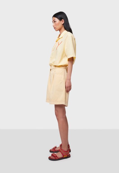 Chemise jaune à manches courtes avec des coutures rouges, associée à un short beige taille haute et des sandales rouges à lanières. Design simple et décontracté.