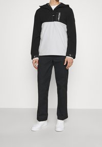 Sportovní bunda v černé a šedé tkanině, se zapínáním na půl zip, kapucí a elastickým páskem. V kombinaci s černými kalhotami a bílými teniskami.