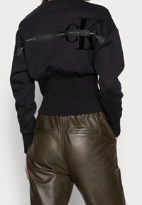 Sudadera negra con banda acanalada, con un gran logo CK en tela mate. Combinada con pantalones de cuero verde oliva con bolsillos traseros.