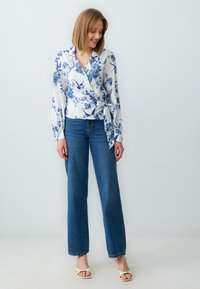 Weiße Wickelbluse mit blauem Blumenmuster, kombiniert mit hoch taillierten blauen Jeans und weißen Blockabsatzsandalen. Glattes Material, lange Ärmel.